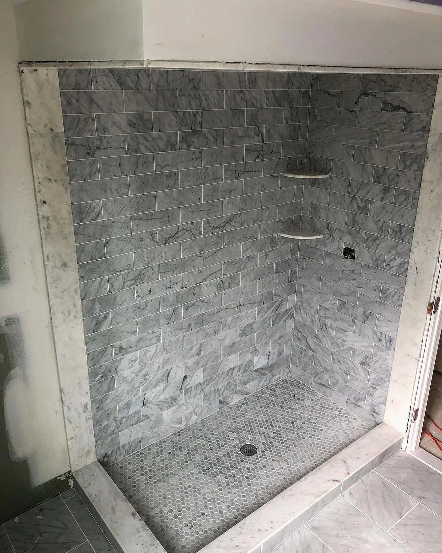 Beautiful gray marble subway tile shower walls with hexagon mosaic floor and corner shelves. by Vicentino Ceramic Tile in Moo