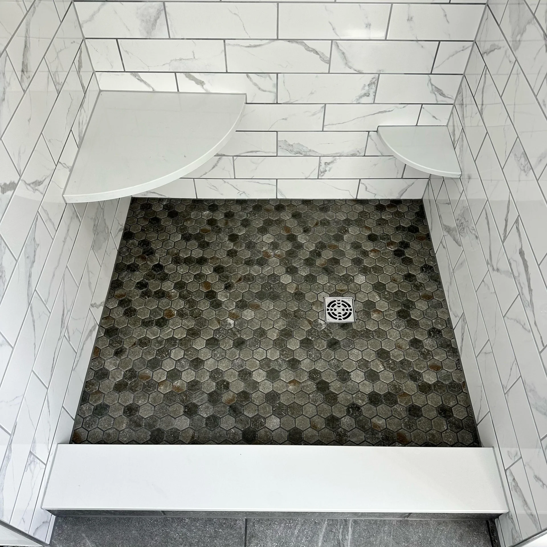 Modern shower with white marble-look subway tiles, dark grey hexagonal floor tiles, and white shelves. by Vicentino Cera