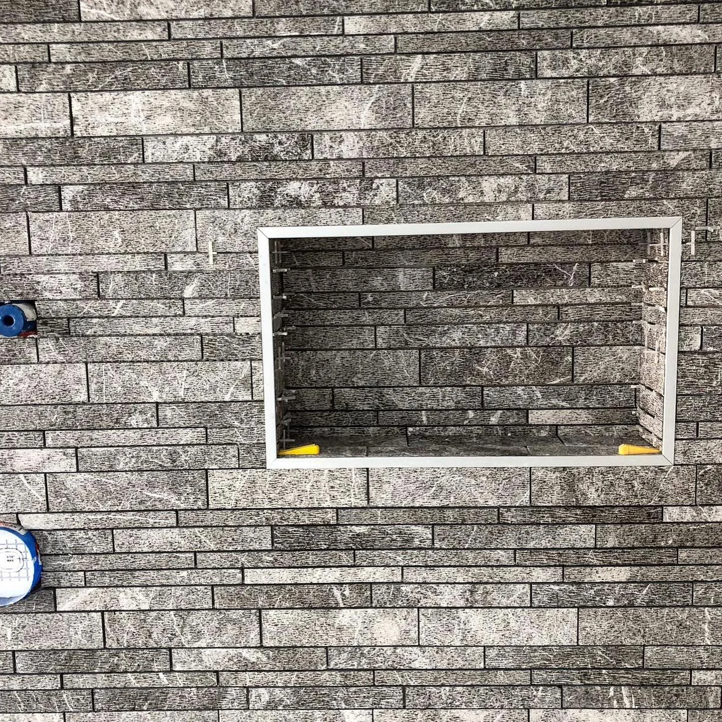 Grey brick-patterned tile wall with a white framed recessed shower niche and plumbing. by Vicentino Ceramic Tile in Yard