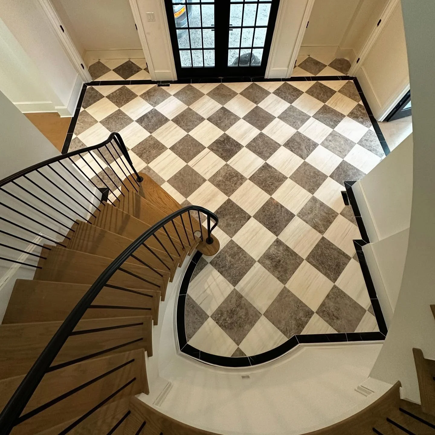 High-angle view of a checkered tile floor with black border and a wooden staircase. by Vicentino Ceramic Tile in Ambler,