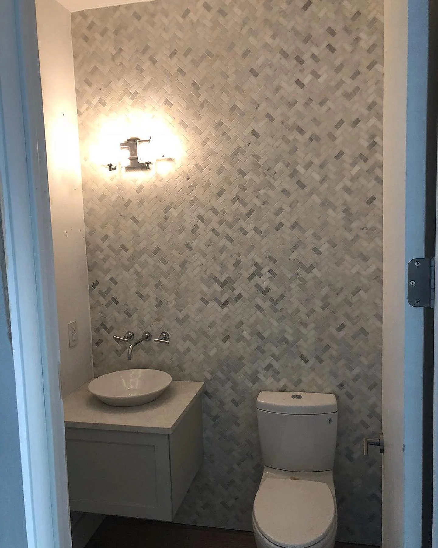 Powder room with light gray herringbone mosaic tile wall, white vessel sink, and toilet. by Vicentino Ceramic Tile in Haddonf