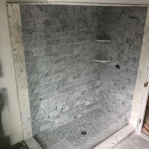 Beautiful gray marble subway tile shower walls with hexagon mosaic floor and corner shelves. by Vicentino Ceramic Tile in Moo
