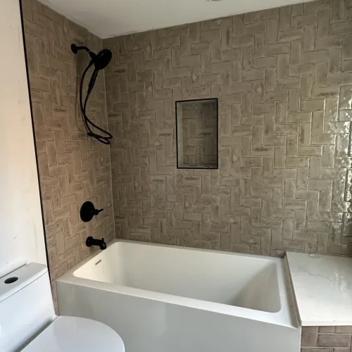 Newly installed bathroom with brown herringbone shower tile, white tub, toilet, and gray floor. by Vicentino Ceramic Tile in