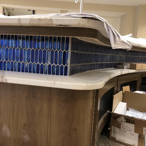 Blue mosaic glass tiles installed on a curved wooden bar with a marble countertop. by Vicentino Ceramic Tile in Huntingdon Va