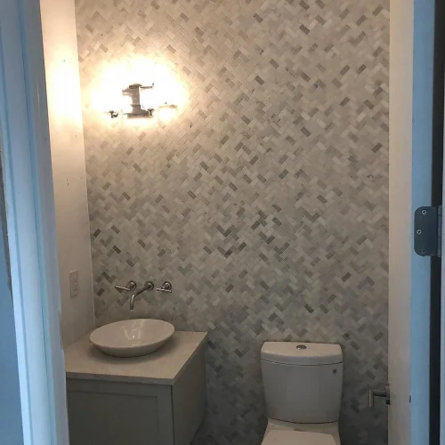 Powder room with light gray herringbone mosaic tile wall, white vessel sink, and toilet. by Vicentino Ceramic Tile in Haddonf
